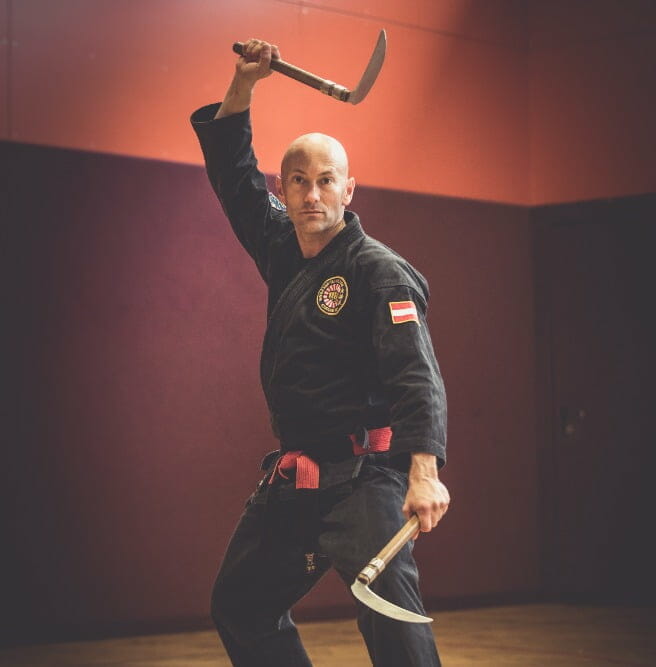 Meisterhafte Verteidigung im Kobudo: In einer Turnhalle nimmt ein Kobudomeister in schwarzem Karateanzug mit rot-schwarzem Gürtel eine Verteidigungsposition ein, bewaffnet mit zwei Kama (kleine Sicheln) in den Händen.