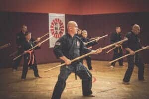 Kraftvolle Praxis einer Kata: In heller Turnhalle mit dem Hintergrund des Sochin Ryu Kobudo Logos stehen zahlreiche Schüler und Meister des Kobudo mit Langstöcken in Angriffsposition. Kobudo in Österreich. Kobudostile. Kobudo trainieren. Kobudo lernen.