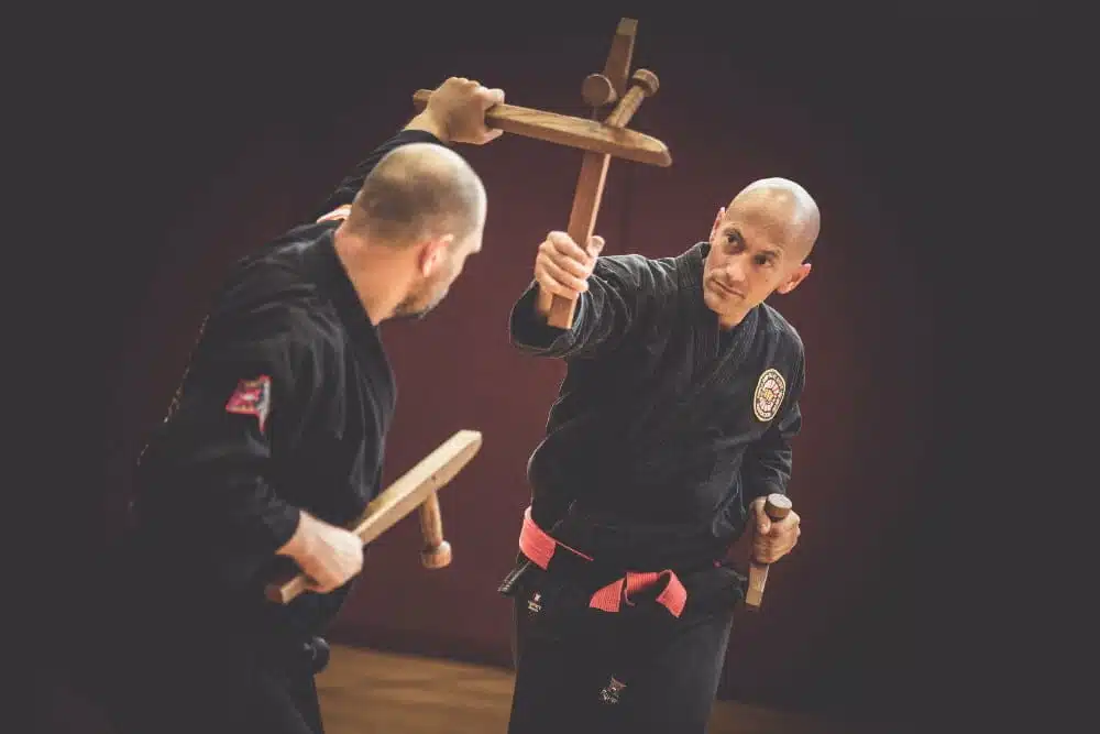 Meisterhafte Choreografie im Kobudo: Zwei Tonfa-bewehrte Meister führen synchronisierten Zweikampf in einer Turnhalle aus. Ein Meister blockiert geschickt den Angriff des anderen. Waffenkampfkunst.