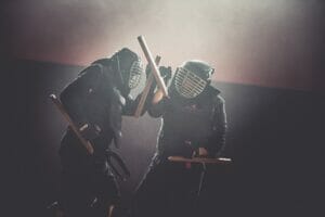 Kampf der Kobudomeister im Vintage-Ambiente: In einer lichtdurchfluteten alten Fabrikshalle führen zwei Meister, ausgerüstet mit Tonfas, einen dynamischen Zweikampf aus. Ein Meister blockiert den Angriff des anderen geschickt.