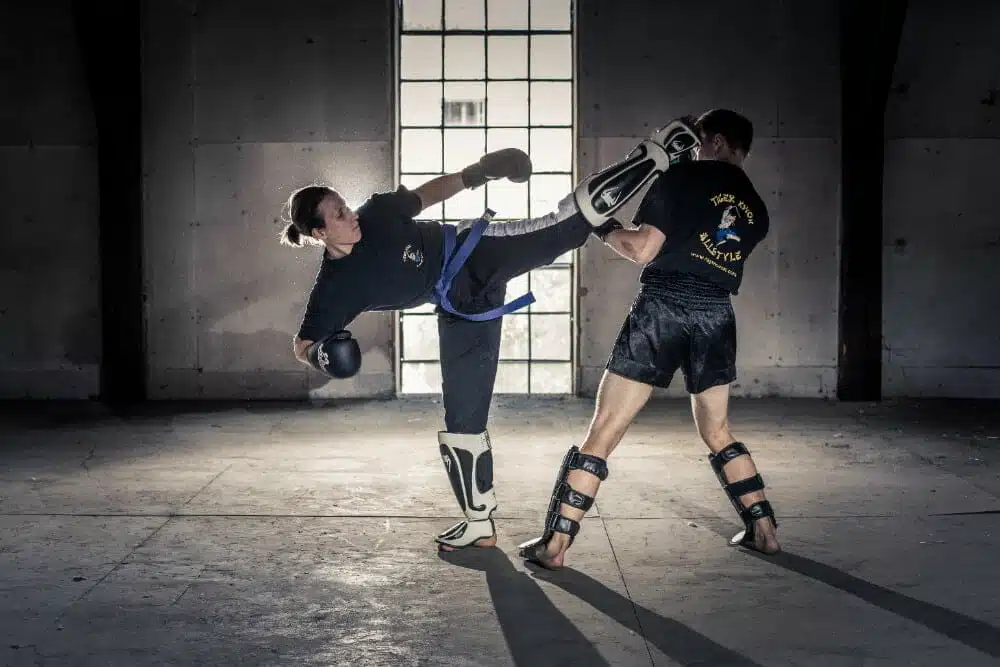 Dynamische Szene im Kickboxen: In einer atmosphärischen alten Fabrikshalle kämpfen eine Kickboxerin und ein Kickboxer in Schutzausrüstung. Durch ein sonnendurchflutetes Fenster im Hintergrund strömt Licht, während die Kickboxerin einen präzisen Halbkreistritt gegen den Kopf des Kickboxers ausführt.