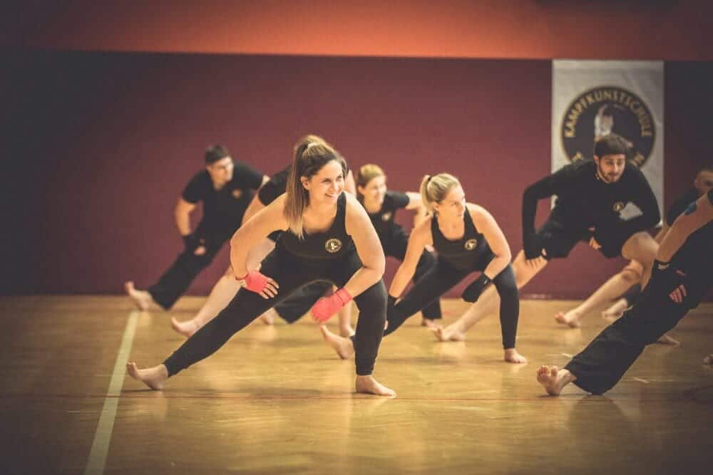 Dynamische Aufwärmung im Kickboxen: Männer und Frauen dehnen sich in einer hellen Turnhalle, mit lächelnden Gesichtern und Blicken zum Kickboxmeister.