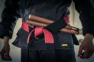 Meisterhafte Präsenz im Kobudo: Mit einem Nunchaku im rot-schwarzen Gürtel steht der Meister in einem Dojo in schwarzem Karateanzug, die Hände entspannt hängend. Das Bild fokussiert auf den Oberkörper und Oberschenkel. schwarze Gürtel, DAN Grade